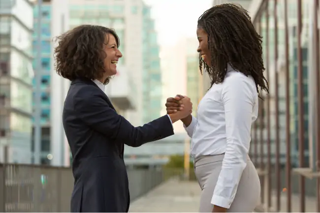 Duas mulheres vestidas com roupas formais sorrindo uma para a outra segurando as suas mãos fechando uma parceria