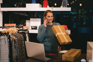 Jovem mulher conversando com um cliente em um smartphone e embalando um produto em uma caixa de papelão para enviar